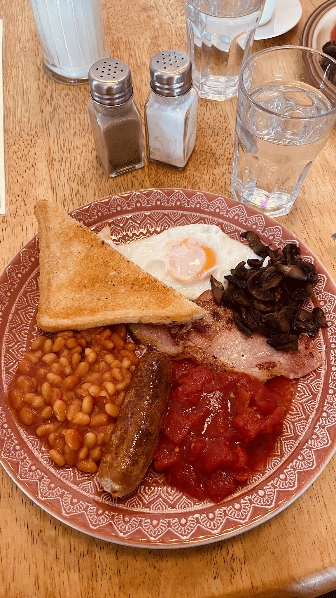 A breakfast on a patterned red plate, with toast, egg, mushrooms, bacon, beans, sausage and tomatoes on a wooden table
