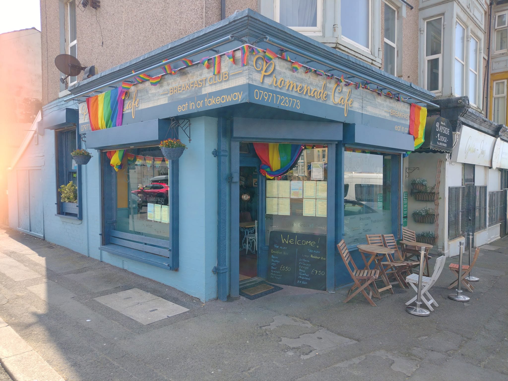 The complete blue corner frontage of Promenade Cafe, including both signs, the café entrance, doorstep, outdoor chairs and pavement